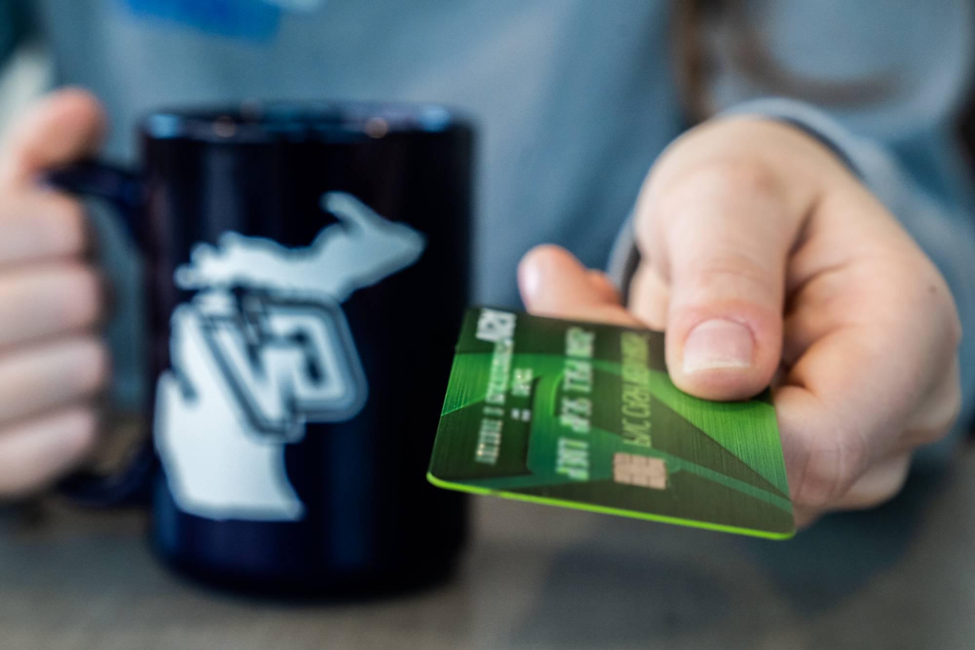 Hand presenting credit card while other hand is holding a GVSU coffee mug